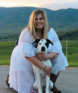 A young blonde woman wearing a white maxi dress hugs a white dog with black facial markings