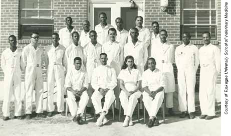 Students in lab coats pose for a black and white photo. There are 6 in the back row, 10 in the middle row, and 4 in the first row, where Alfreda Webb is seated. There is a brick building in the background.